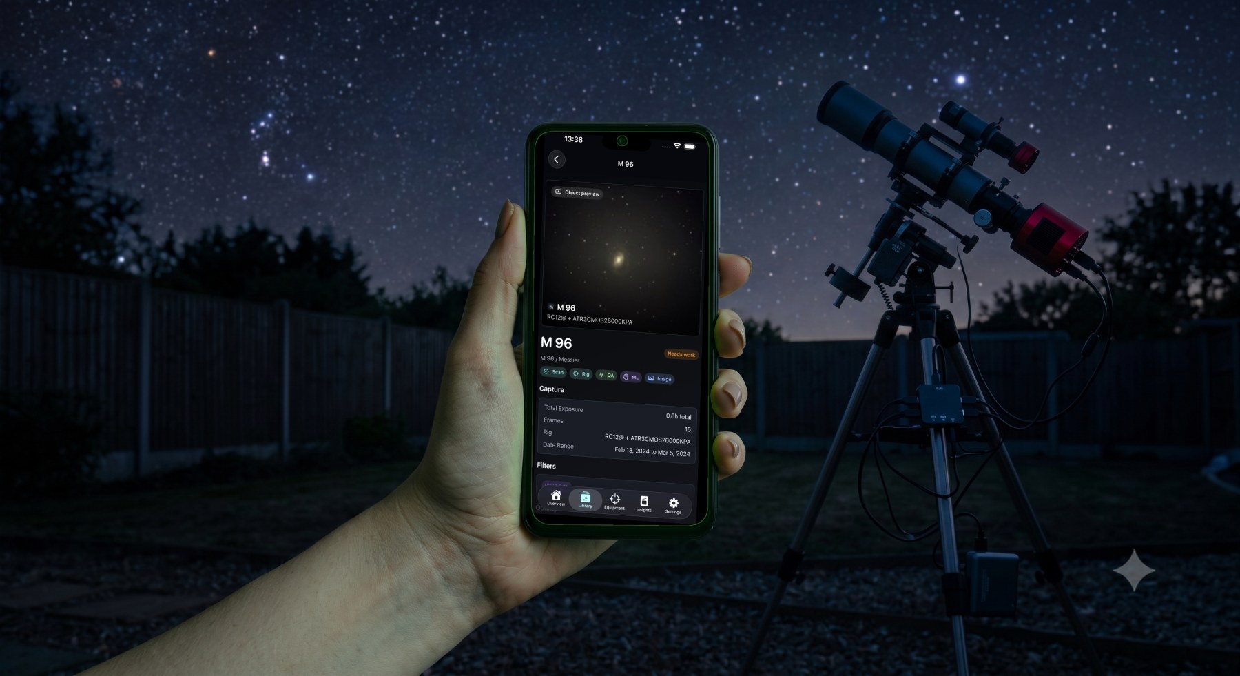 A hand holding a phone running the AstroIndexer companion app at night, showing the M 96 object page, with a telescope rig under a starry sky in the background